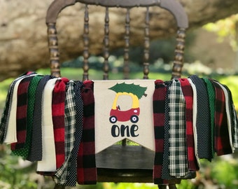 Puede incluir: Una festiva pancarta para trona con un diseño de árbol de Navidad y coche, perfecta para una celebración de primer cumpleaños. La pancarta presenta una mezcla de tiras de tela rojas, verdes, negras y blancas, y la palabra "one".