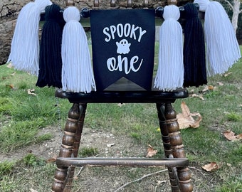 Op de afbeelding: Een zwart-witte banner met de tekst "Spooky one" en een spookafbeelding. De banner is bevestigd aan een houten hoge stoel met zwarte en witte garenkwastjes.