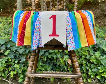 May include: A wooden high chair decorated with a colorful banner. The banner has a neutral background with a large red number 1. The chair is surrounded by greenery.