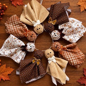 May include: A circular arrangement of seven fabric ghost ornaments featuring various patterns including brown plaid, tan polka dots, dark brown geometric prints, and white floral designs. Each ghost has two black bead eyes and a small velvet bow.