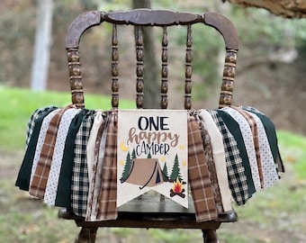 May include: A fabric banner featuring a central pennant with the text ONE happy CAMPER, a tent, pine trees, and a campfire graphic. The banner is decorated with alternating strips of fabric in brown, dark green, and white with plaid or dot patterns.