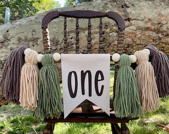 Pode incluir: Uma faixa branca com a palavra "one" em letras pretas pendura em uma cadeira de madeira. A faixa é decorada com uma guirlanda de borlas de fio marrom, bege e verde.