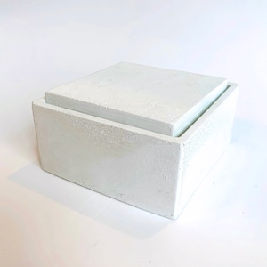 Pode incluir: Uma caixa quadrada de concreto branco com uma tampa. A caixa tem uma superf&iacute;cie &aacute;spera e texturizada.