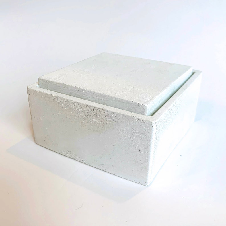 Pode incluir: Uma caixa quadrada de concreto branco com uma tampa. A caixa tem uma superf&iacute;cie &aacute;spera e texturizada.