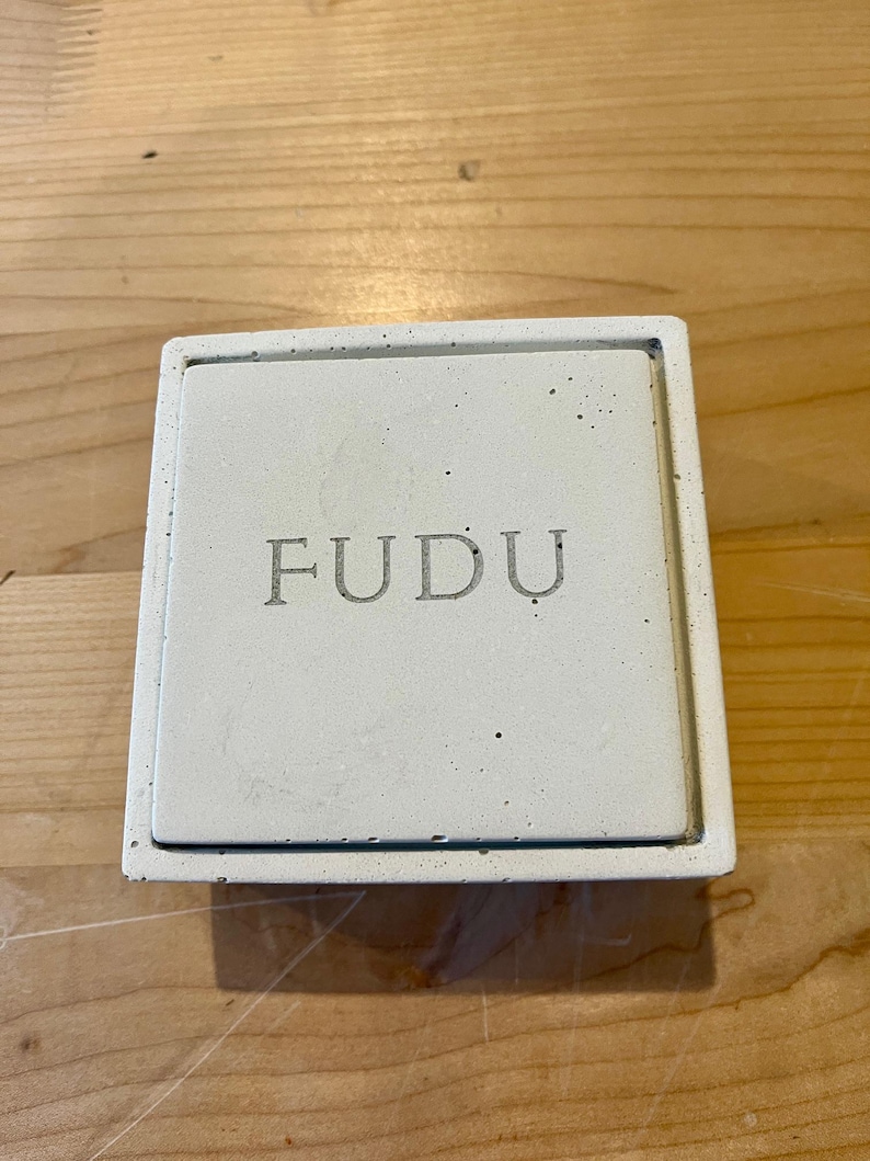 Pode incluir: Uma caixa quadrada branca com a palavra "FUDDU" em relevo na parte superior.
