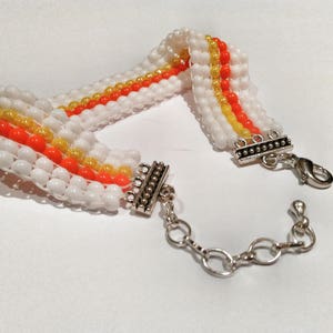 May include: A beaded bracelet featuring white, orange, and yellow beads. The bracelet has a silver-toned clasp and chain. The beads are arranged in horizontal rows, creating a striped pattern. The bracelet is designed to be worn around the wrist.