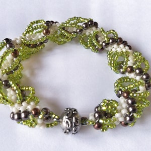 Op de afbeelding: Een kralenarmband met een herhalend patroon van groene, witte en bruine kralen. De armband heeft een zilverkleurige sluiting. De kralen zijn gerangschikt in een bloemen- of bladvormig ontwerp, waardoor een textuur ontstaat.