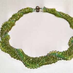 Op de afbeelding: Een groene kralenketting met een spiraalvormig ontwerp. De ketting is gemaakt van kleine, ronde kralen en heeft een zilverkleurige sluiting. De kralen variëren in kleur, wat een textuur creëert. Ongeveer 40 cm lang.