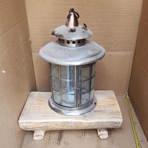 May include: A silver lantern with a glass paneled design sits on a rustic wooden base. The lantern has a copper colored top and a handle.