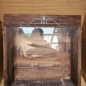 May include: A dark brown wooden shadow box with a glass front. The box has a decorative scroll design on the top and the words "Treasured Memories" inscribed on the bottom.