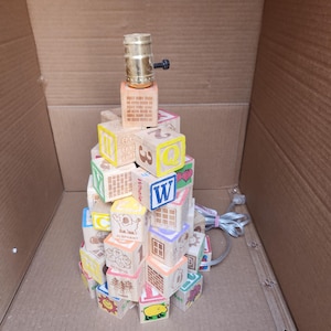 May include: A whimsical lamp made from stacked wooden alphabet blocks. The blocks feature colorful letters, numbers, and illustrations. The lamp is topped with a brass lamp holder and a black switch. The lamp is approximately 12 inches tall.