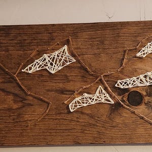 May include: A wooden wall hanging with a white string art design of mountains. The mountains are made of white string stretched between nails hammered into the wood.