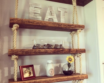 May include: Three wooden shelves with rope hangers, the top shelf has the word "EAT" in white letters, the middle shelf has a glass container with small rocks, and the bottom shelf has a glass vase with a sunflower, a glass bottle, and a framed picture.