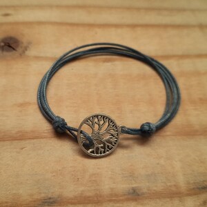 May include: A bracelet with a silver-coloured tree of life charm. The bracelet has a dark grey cord, knotted at each end. The charm is circular with an intricate tree design. The bracelet is displayed on a wooden surface.