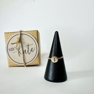 Può includere: Un anello color oro con un design a punto e virgola è esposto su un espositore nero a forma di cono. Una piccola scatola regalo quadrata con un'etichetta rotonda che recita "made by kate" è legata con spago.