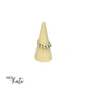 Peut inclure: Une bague argentée ornée de plusieurs petites perles rondes est présentée sur un présentoir conique beige. La bague, au design simple et élégant, est mise en valeur. Le fond blanc uni met en évidence les détails de la bague.