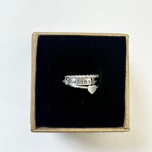 Op de afbeelding: Een set van drie zilveren ringen in een kleine doos. Eén ring is gedraaid, een andere heeft de naam "Savannah" gegraveerd, en de derde heeft een kleine hartvormige bedel. De ringen worden gepresenteerd tegen een zwarte fluwelen achtergrond.