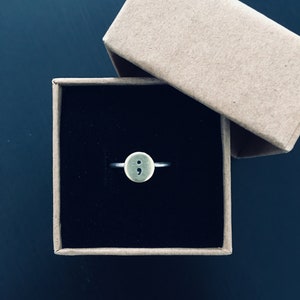 Pode incluir: Um anel de prata com uma face circular dourada com um símbolo de ponto e vírgula. O anel é apresentado dentro de uma pequena caixa de presente quadrada de cartão aberta, com um interior preto, sobre um fundo escuro.