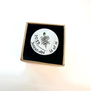 May include: A silver-colored heart-shaped pendant with a medical symbol and a small green gem. The pendant is engraved with "2025 Jordan, RN U of M" and is presented in a small, open cardboard box with a black interior.
