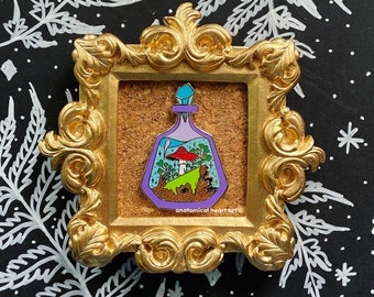 May include: A gold-framed picture with a purple glass bottle containing a miniature forest scene with a red mushroom, green trees, and brown rocks. The bottle has a blue crystal stopper and the text "anatomical heart art ©" is printed below the bottle.