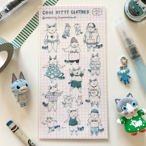 以下が含まれることがあります： 「COOL KITTY CLOTHES」と題されたステッカーシート。様々な衣装を着た猫のイラストが描かれています。猫は青と白で、パーカー、帽子、ドレスを着ています。その他、青いフィギュア、クレヨン、マスキングテープなどがあります。