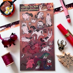 May include: A sheet of stickers with cartoon cats in various poses, some with demonic features. The sheet includes the text "UNNATURAL EVIL BEING" and the artist's social media handle. The stickers are colourful and whimsical.