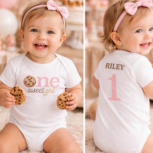 Peut inclure: Body bébé blanc avec l'inscription "one sweet girl" et un motif de biscuit aux pépites de chocolat. Le dos du body porte le nom "Riley" et le chiffre "1". Le bébé porte un bandeau rose et tient deux biscuits.