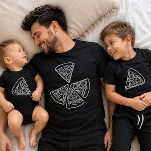 May include: Black t-shirts and a onesie with a white pizza slice design. The adult shirt features four pizza slices arranged in a circle. A baby and a child are wearing matching shirts, and the family is lying on a bed.