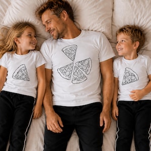 Peut inclure: Trois personnes portant des t-shirts blancs avec des motifs de tranches de pizza noires. Les chemises sont associées à des pantalons noirs. Les motifs de tranches de pizza sont en noir et blanc. Ils sont allongés sur un lit blanc.