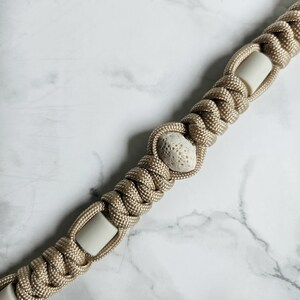 May include: A tan paracord lanyard featuring a series of woven knots interspersed with smooth, cylindrical off-white beads and a single, central, porous, textured off-white stone bead, creating a repeating pattern along the length of the cord.