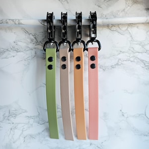 Puede incluir: Cuatro correas de cuero para la muñeca en colores pastel: verde, beige, melocotón y rosa. Cada correa tiene herrajes negros y dos remaches negros. Las correas cuelgan sobre un fondo de mármol blanco y gris.