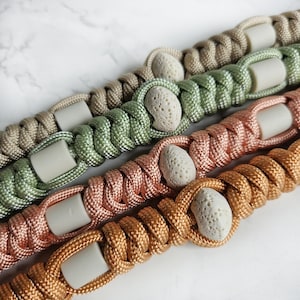 Op de afbeelding: Vier gevlochten touw armbanden met natuurlijke stenen kralen. De armbanden zijn in de kleuren beige, groen, roze en bruin. Elke armband heeft een andere kleur en heeft een witte stenen kraal.