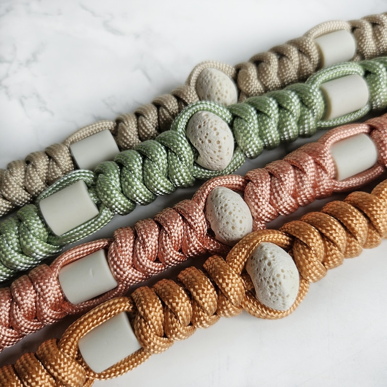 May include: Four braided paracord lanyards in tan, sage green, rose gold, and copper. Each lanyard features a repeating pattern of smooth, cylindrical, light gray beads alternating with textured, porous, off-white lava stone beads.