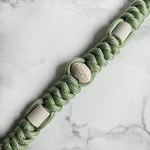 May include: A sage green paracord lanyard featuring a series of intricate knots. The design incorporates smooth, cylindrical off-white beads alternating with a single, porous, textured, light beige stone bead centered within the woven cord pattern.