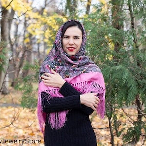 May include: A woman wearing a pink and black patterned headscarf with fringe. The scarf is wrapped around her head and neck. She is standing in a forest with fall foliage.