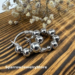 May include: Silver hoop earrings with multiple round beads. The earrings are displayed on a wooden surface with a natural grain pattern. Delicate white flowers are placed near the earrings.