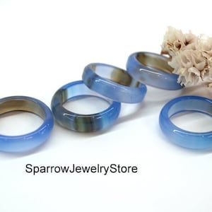 Pode incluir: Cinco anéis de ágata azul com um padrão de faixas brancas e marrons. Os anéis estão empilhados uns sobre os outros. Os anéis estão sobre uma superfície branca com um ramo de flores brancas.