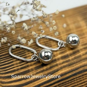 Puede incluir: Pendientes de aro plateados con un dije esférico colgante. Se exhiben sobre una superficie de madera, con delicadas flores blancas en el fondo. Los pendientes tienen un acabado pulido y reflectante.