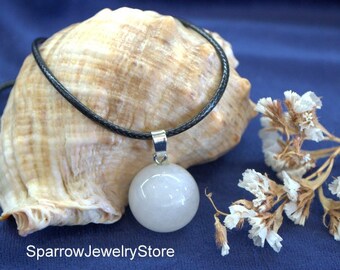 May include: A white gemstone pendant necklace on a black leather cord. The pendant is a round, smooth sphere. The necklace is displayed on a seashell and a sprig of dried white flowers.