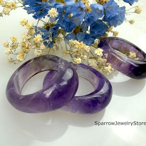 Op de afbeelding: Drie gladde, gepolijste amethist ringen met dieppaarse tinten en natuurlijke kleurvariaties. De ringen zijn in een groepje gerangschikt en tonen hun afgeronde, dikke bandontwerp. De tekst SparrowJewelryStore is zichtbaar in de rechterbenedenhoek.