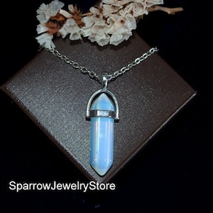 Puede incluir: Un collar de cadena de plata con un colgante de cristal iridiscente azul claro en forma de punta. El colgante está engastado en un bisel de plata y tiene un anillo de plata alrededor de la parte superior. El collar se muestra en una caja de terciopelo marrón. El texto "SparrowJewelryStore" es visible en la parte inferior de la imagen.