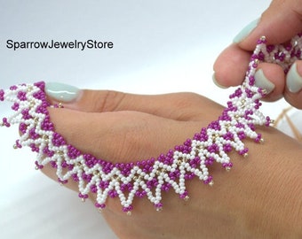May include: A white and purple beaded necklace with a zig-zag pattern. The necklace is made with small seed beads and has a delicate, feminine look.
