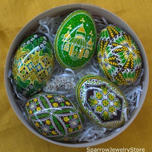 Pode incluir: Uma coleção de cinco ovos de Páscoa decorados de forma vibrante numa caixa redonda de madeira cheia de papel branco picado. Os ovos apresentam padrões intrincados em verde, amarelo e castanho, com desenhos florais e geométricos. Artesanato tradicional da Páscoa.