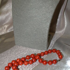 Peut inclure: Un collier de perles de corail rouge avec un fermoir en argent. Le collier est pos&eacute; sur une surface en tissu blanc.
