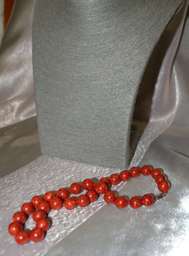 Peut inclure: Un collier de perles de corail rouge avec un fermoir en argent. Le collier est pos&eacute; sur une surface en tissu blanc.