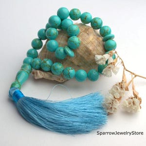 May include: Turquoise blue gemstone prayer beads with a blue tassel, resting on a seashell with dried white flowers.