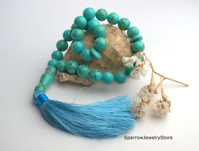 May include: Turquoise blue gemstone prayer beads with a blue tassel, resting on a seashell with dried white flowers.