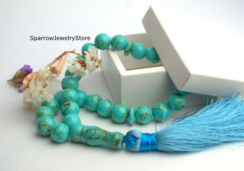 May include: A turquoise blue prayer bead necklace with a light blue tassel. The necklace is in a white box with the text "SparrowJewelryStore" on the box.