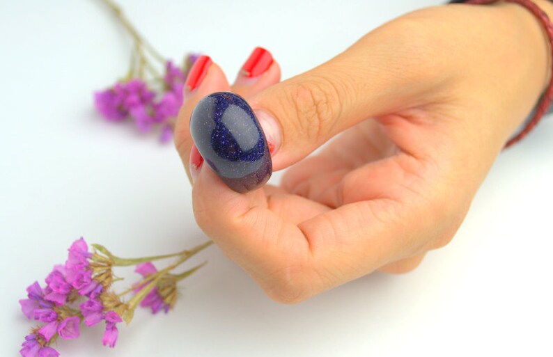 May include: A dark blue, oval-shaped gemstone with a glittery finish. The gemstone is being held by a hand with red nail polish.