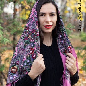 May include: A pink patterned headscarf with floral designs and fringe. The scarf is worn by a person with long dark hair and red lipstick.
